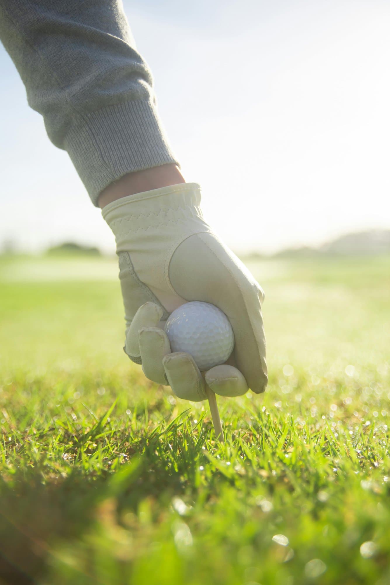 Hand i vit golfhandske som sätter upp en golfboll på en peg i gräset, i motljus